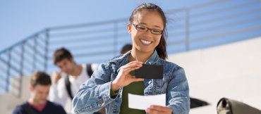 Image of a woman taking a picture of a check while sitting outdoors on a type of bleacher.