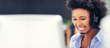 Woman in a blue button down shirt with a head-set on, smiling while looking at a computer monitor.