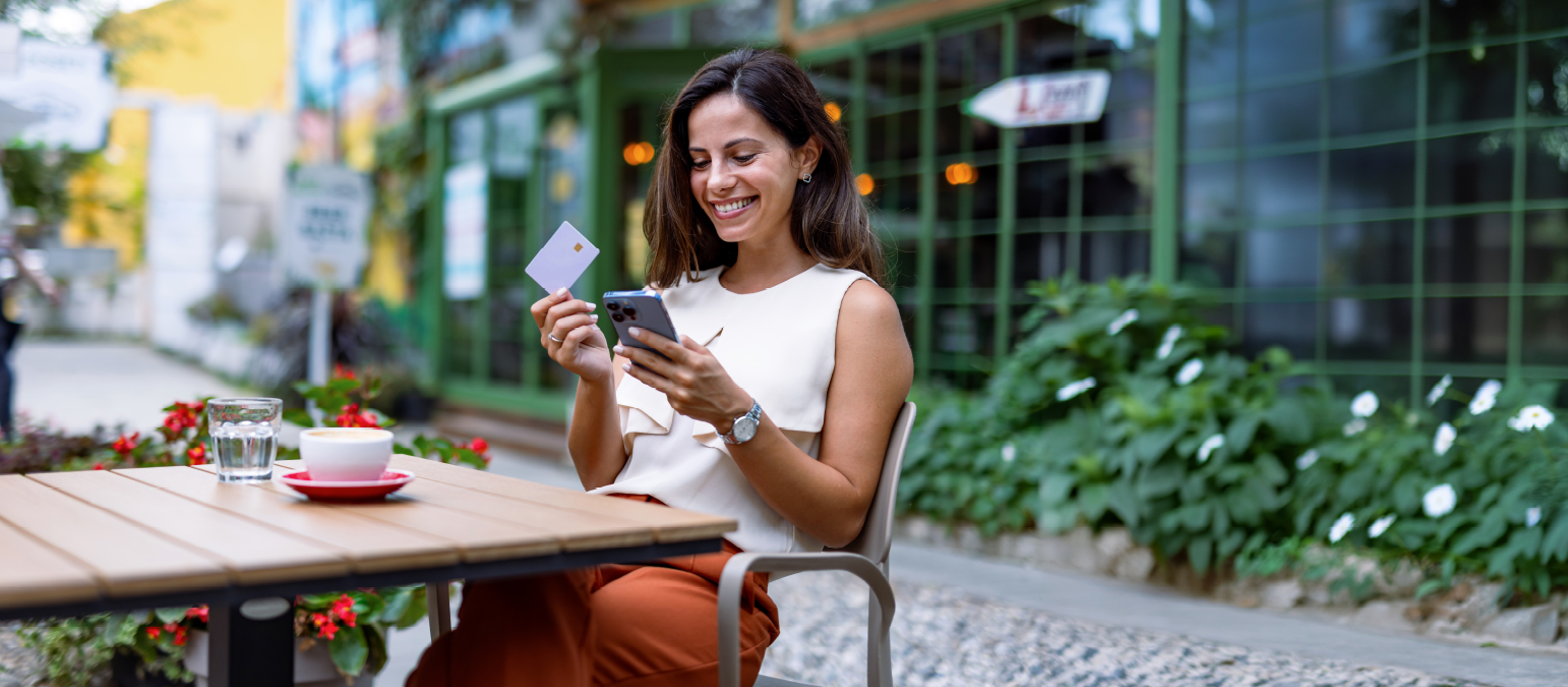 Image of a woman sitting an a table outdoors. She is holding a plastic card in one hand and a cell phone in the other.