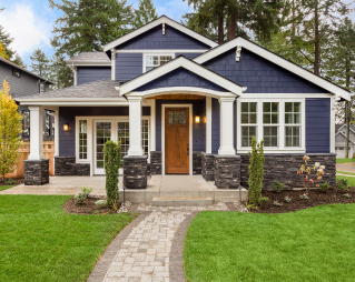 Photo of craftsman style house, front view.  House is medium blue color with white trim, and a stained front door.  Home is surrounded by green grass and a stone walkway leading to the porch.