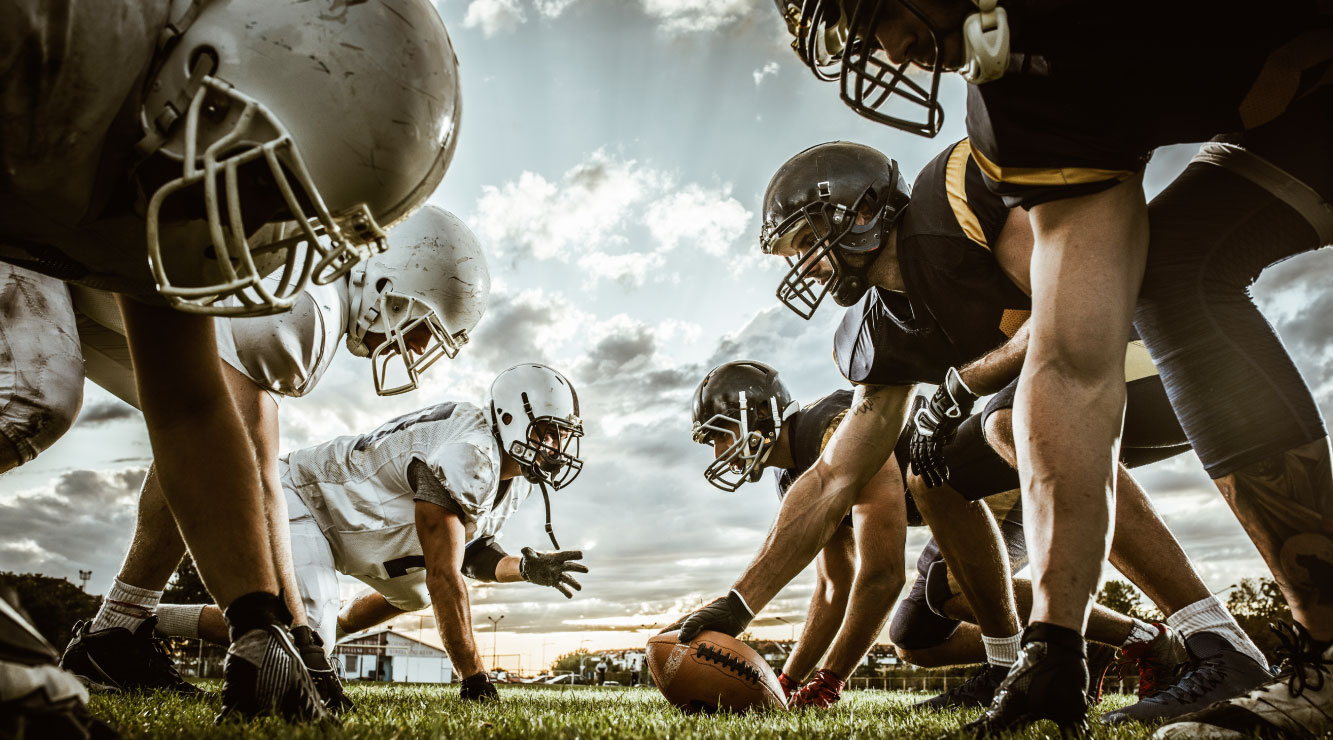 Image of football players head-to-head preparing for a play on a green football field with a lightly clouded sky in the background.  One team has white uniforms and helmets, the other team has black uniforms and helmets.
