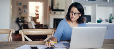 Image of a woman wearing glasses sitting her in kitchen in front of a laptop, also using her mobile phone, with papers spread around the table.