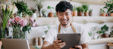 Image of a man working at a floral shop with a laptop in front of him, and he's holding a portable electronic device.