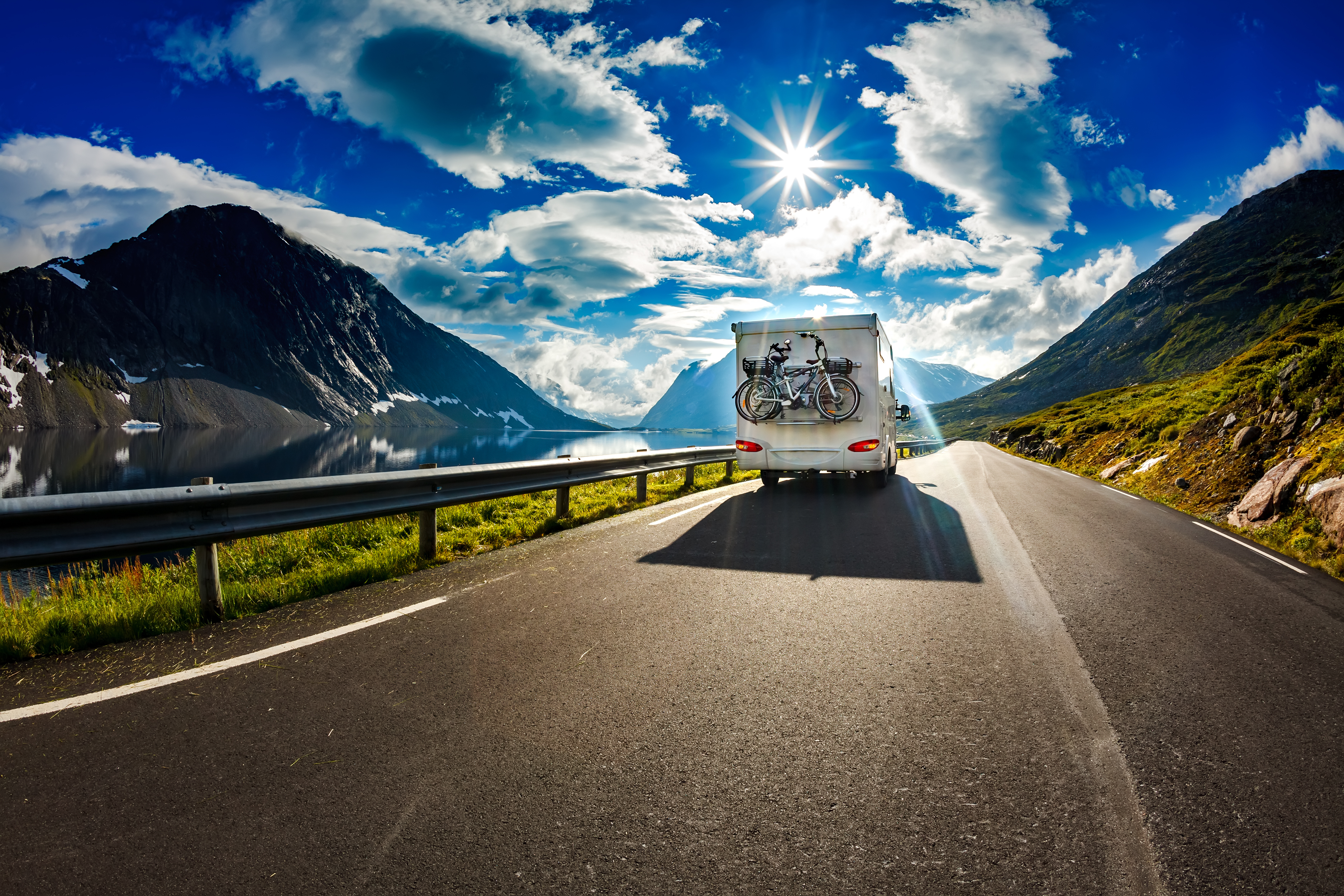 RV with bicycles on back driving down a road winding between two mountains on a sunny day with scattered clouds.