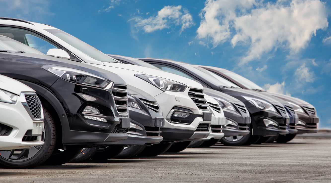 Image of a row of cars on concrete surface.  Blue sky with scattered white clouds in the background.