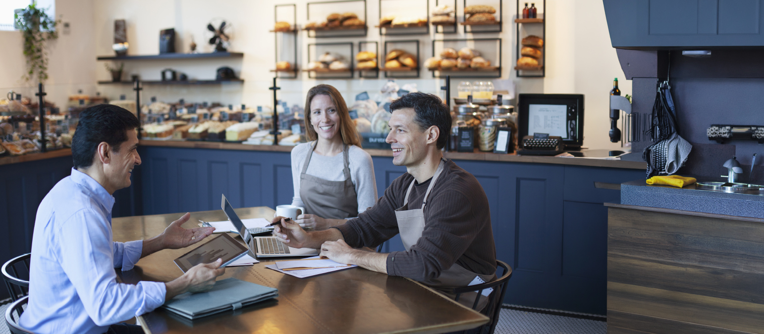 Image of 3 individuals in a cafe setting. Two have aprons and the other is sitting across the table from them.