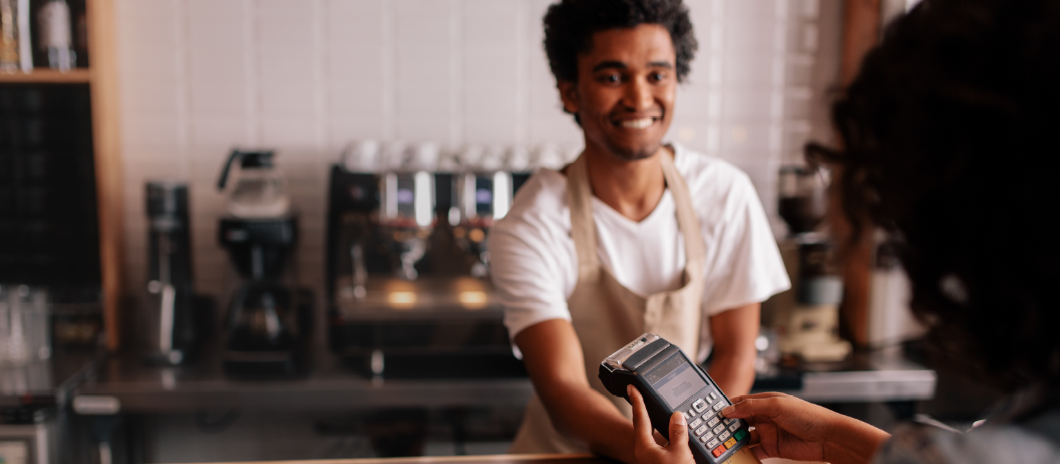 Image of a man working at a coffee shop, assisting a woman in paying with her card using a portable card scanning device.