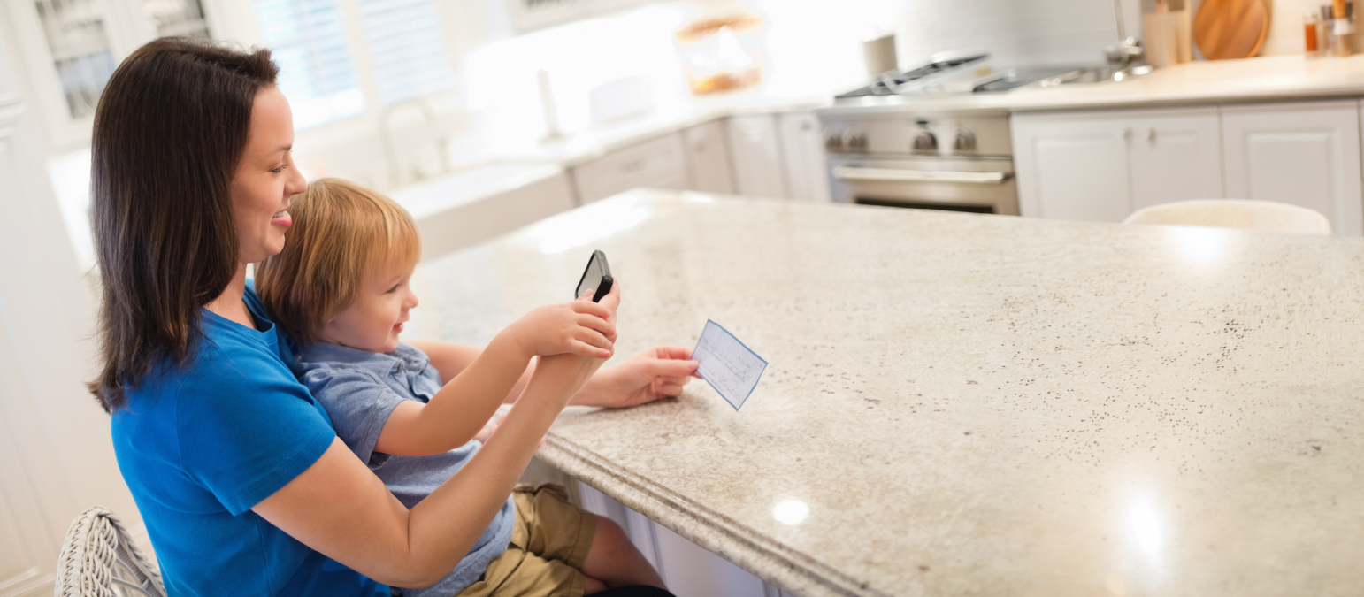 Image of a child sitting on a woman's lap, photo taken from the side. The two are taking a photo of a slip of paper in a kitchen setting.