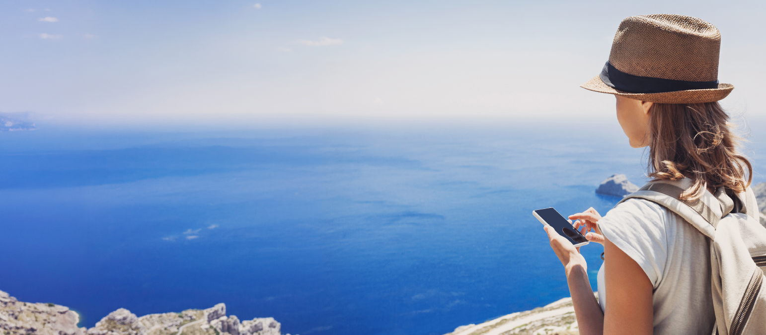 Image from the back corner of a woman holding a cell phone standing in front of an ocean. She has on a hat and backpack.