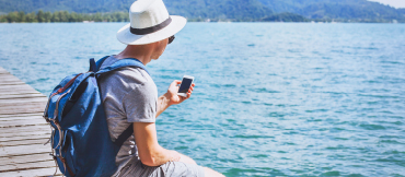 Image of a person sitting on a dock with a blue backpack on.  The doc is surrounded by water with mountains in the background.  The individual has a white hat on and is looking at their mobile device.
