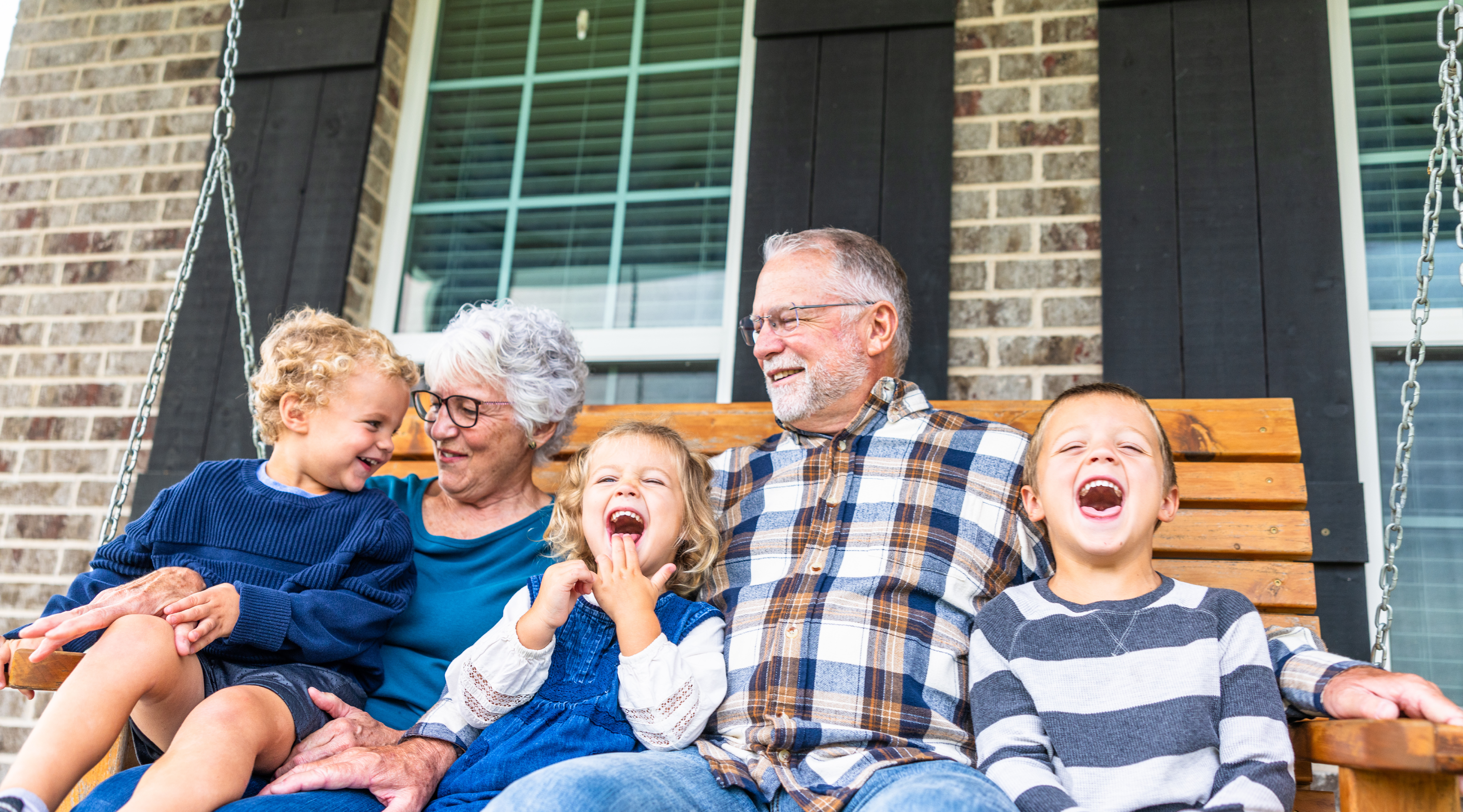 Image of 3 young children and two adults sitting on a wooden porch swing in front of a brick house with windows and black shutters.  All individuals appear to be smiling and happy.