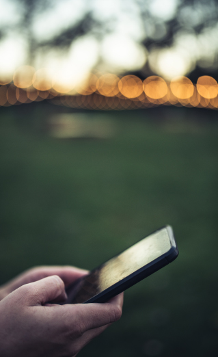 Set of hands holding a mobile phone.  View is from the side of the hands and phone.  Background appears to be blurred out green grass with a sunset and glowing lights.